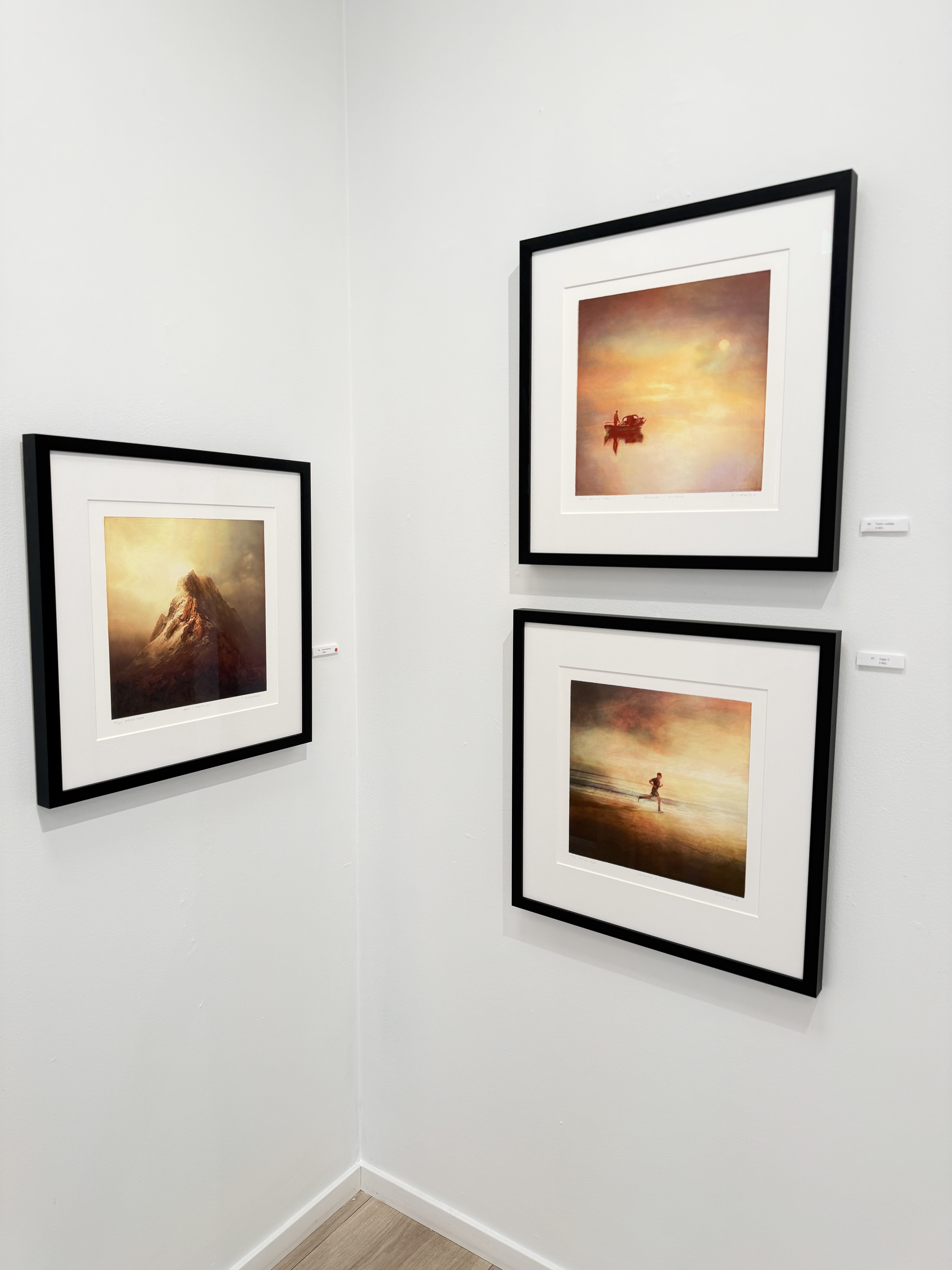 Tre innrammede motiver i varme farger: et fjell, en båt og en løper, fotografert i et hjørne av galleriet.