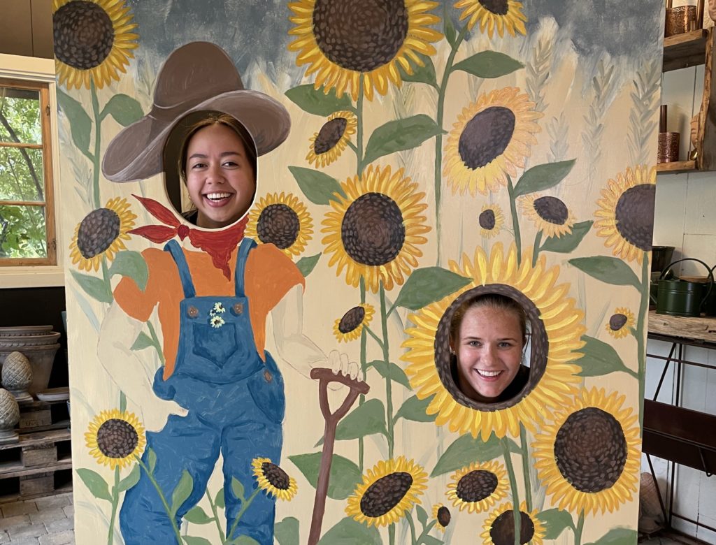 Maria og Solfrid, ansatte på Lundbergs Planteskole, i en håndmalt bildevegg med gartner og solsikker