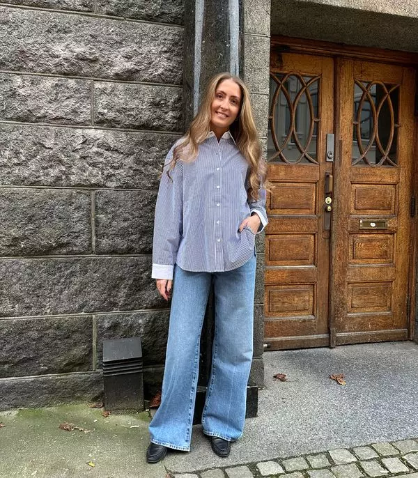 Jente står faen en vegg med brun dør viser frem en stripete skjorte, til har hun lyse jeans og svart sko