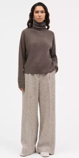 Jente med svart hår står med en brun høyhalset genser med beige bukser til