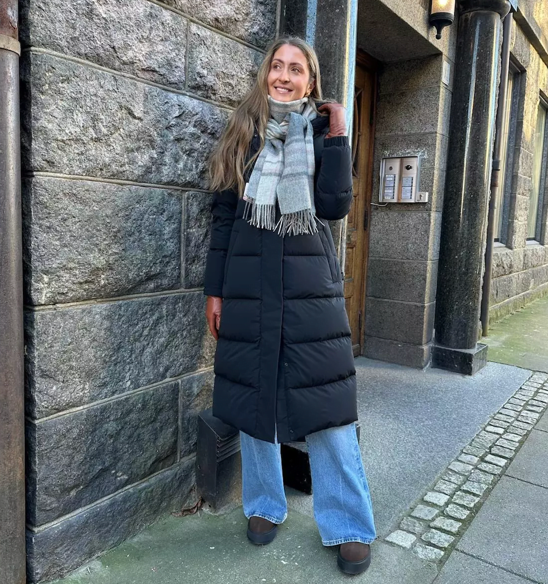 Jente i lang dunkåpe fra Woolrich med ett rutete skjerf i grå toner og lyse jeans bukser. Brune skinnhansker og boots.